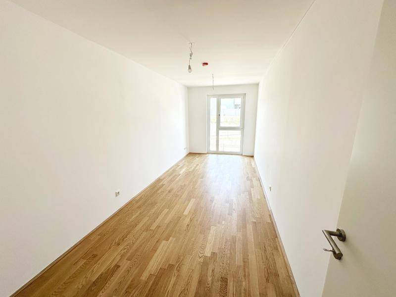 Helles, leeres Zimmer mit Holzboden und einer Glastür, die nach draußen führt, in einem Neubau.