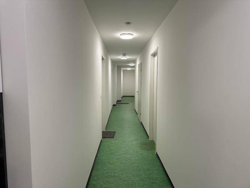 Langer, heller Flur mit weißen Wänden und durchgehend grünem Linoleumboden.