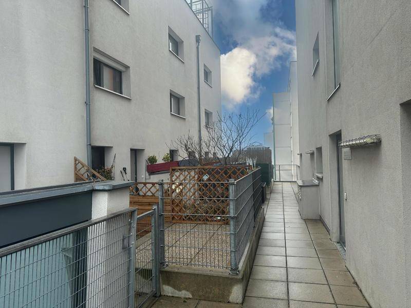 Gepflasterter Außenbereich mit Terrasse und Zugangsweg zwischen Gebäuden, unter blauem Himmel.