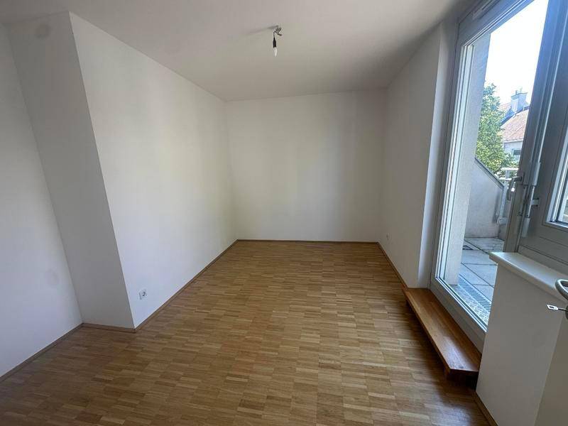 Leeres Zimmer mit Parkettboden und großem Fenster, das Zugang zu einem Außenbereich bietet.