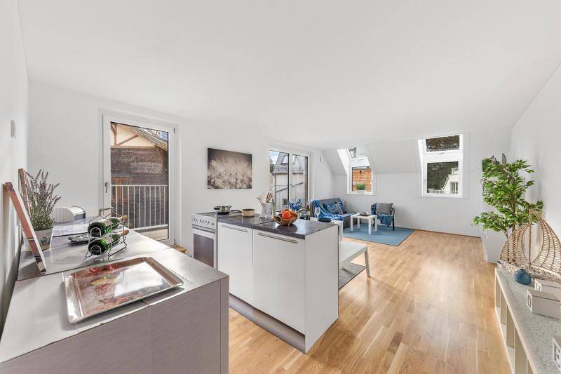 Heller offener Wohnbereich mit kleiner Küche, Holzboden und Zugang zu einem Balkon.