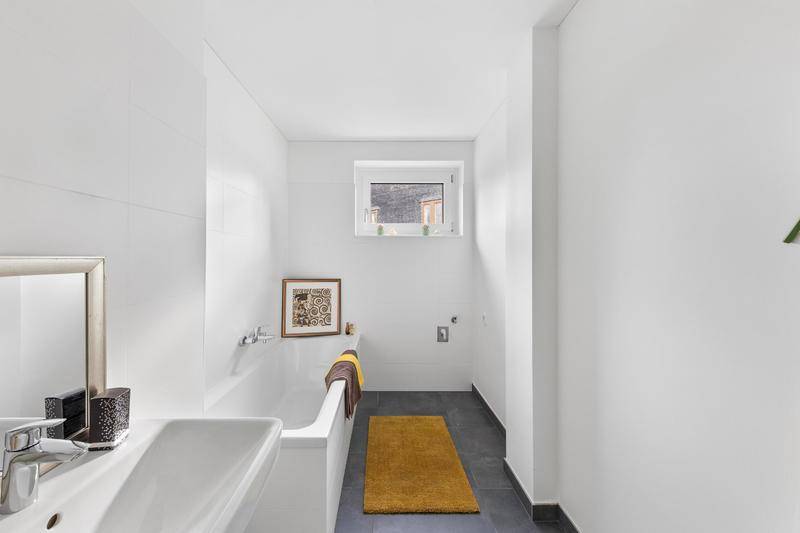 Funktionales Badezimmer mit Badewanne, Einzelwaschbecken, Fenster und grauen Bodenfliesen.