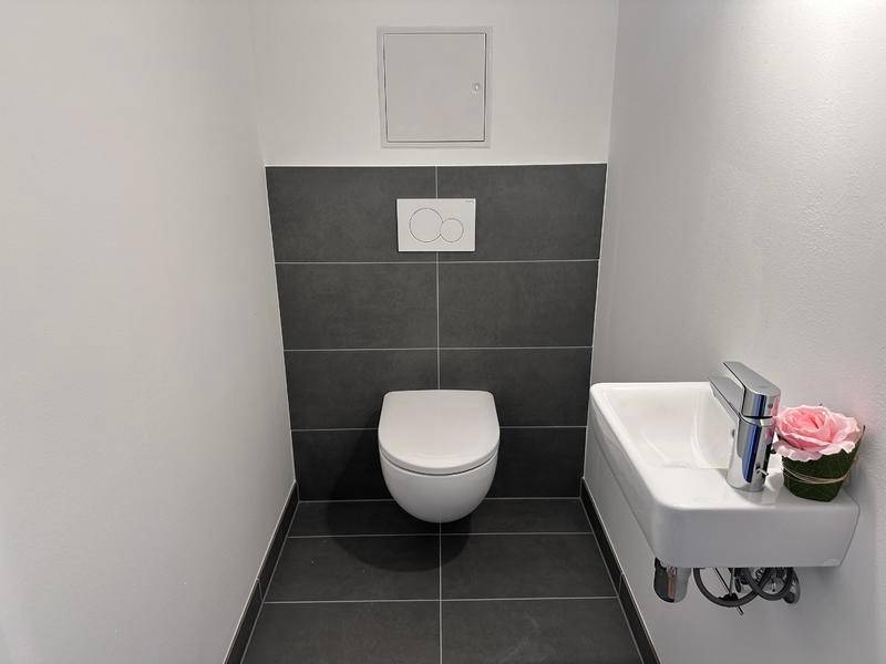 Gäste-WC mit wandhängender Toilette, kleinem Waschbecken und modernen grauen Wandfliesen.