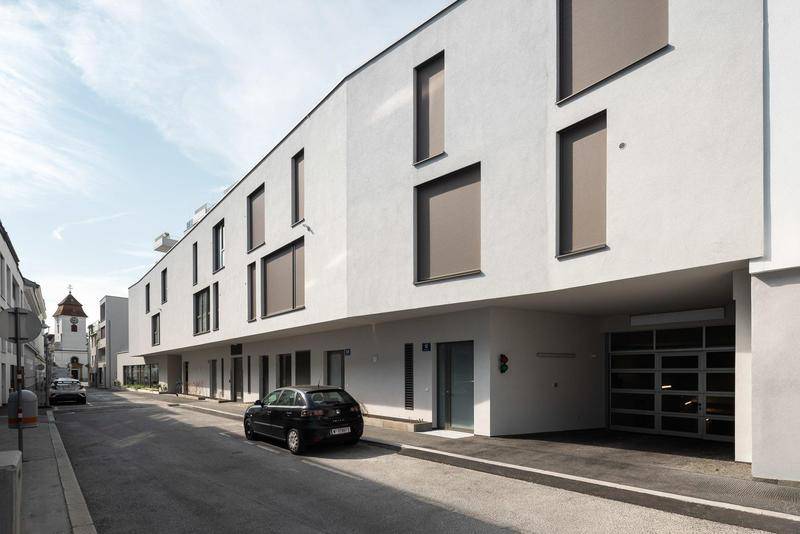 Moderne Gebäudefassade mit großen Fenstern und einem Eingang zur Tiefgarage in einer Seitenstraße.