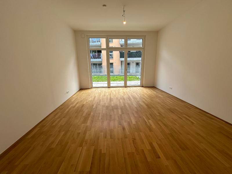 Helles, leeres Zimmer mit Holzboden und großen Fenstern zum Gartenbereich.