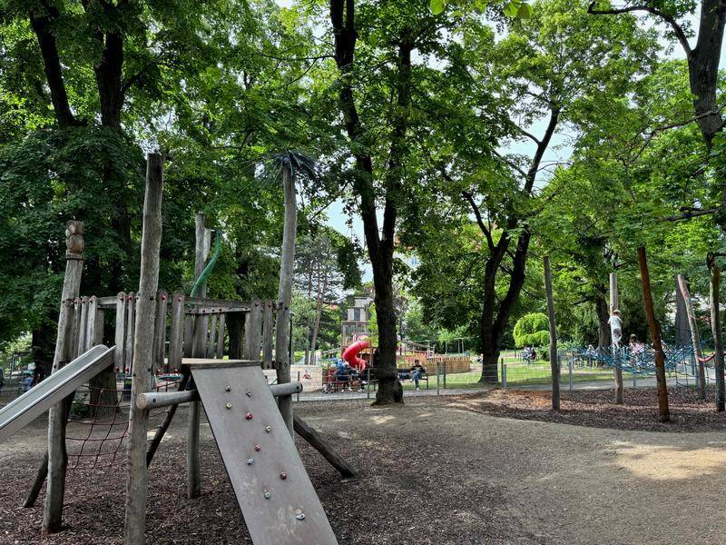Großer Kinderspielplatz mit verschiedenen Holzspielgeräten, umgeben von hohen Bäumen.