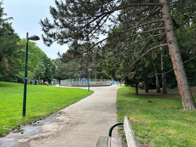 Spazierweg durch eine grüne Parkanlage mit Bäumen, Bänken und einem entfernten Spielplatz.