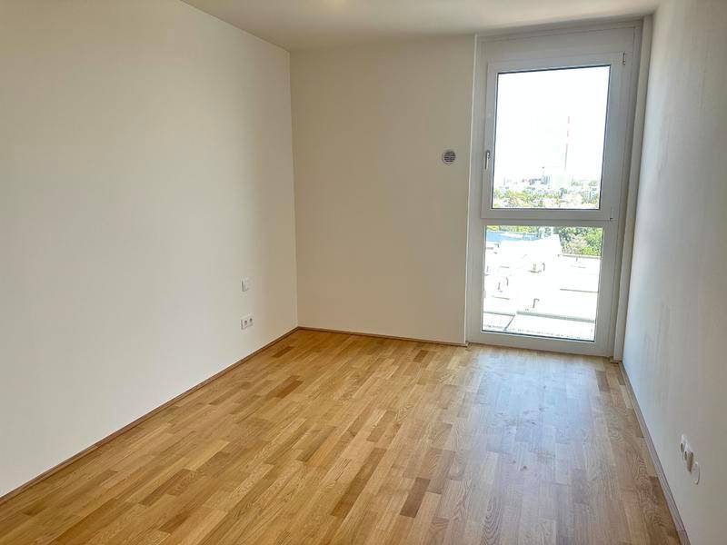 Leeres Zimmer mit Holzboden und großem Fenster, das viel Tageslicht hereinlässt.