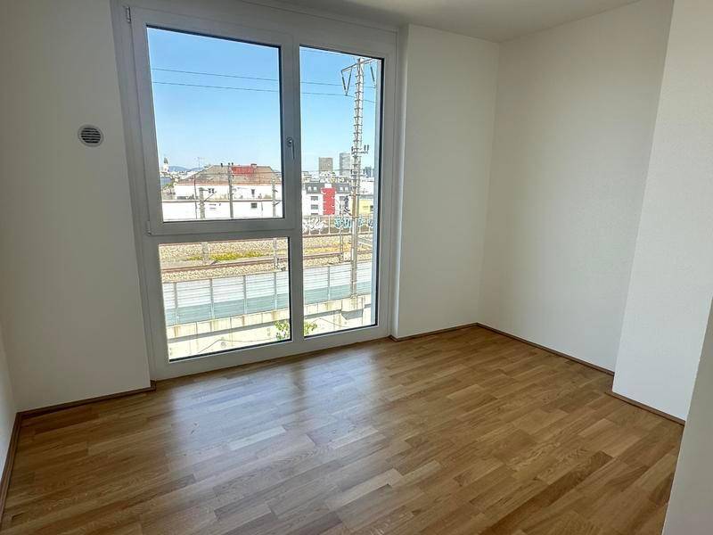 Leeres Zimmer mit Holzboden und großem Fenster, das einen Blick auf die Bahngleise bietet.