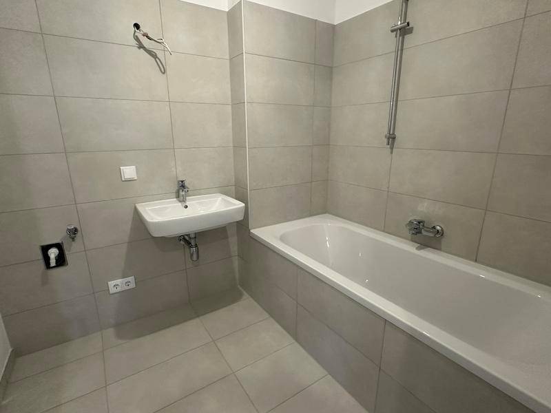 Modernes Badezimmer mit hellgrauen Fliesen, einer Badewanne mit Duschfunktion und einem Waschbecken.