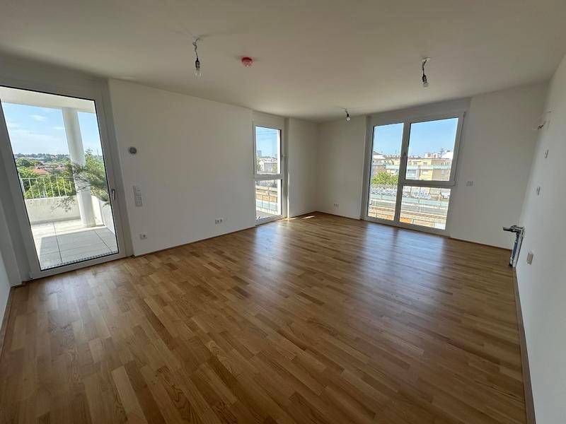 Helles, leeres Wohnzimmer mit Holzboden und großen Fenstern, die viel Tageslicht hereinlassen.