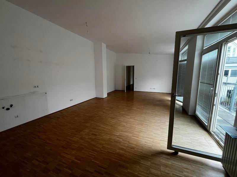 Heller, leerer Raum mit Parkettboden und breiter Fensterfront, die viel Tageslicht hereinlässt.