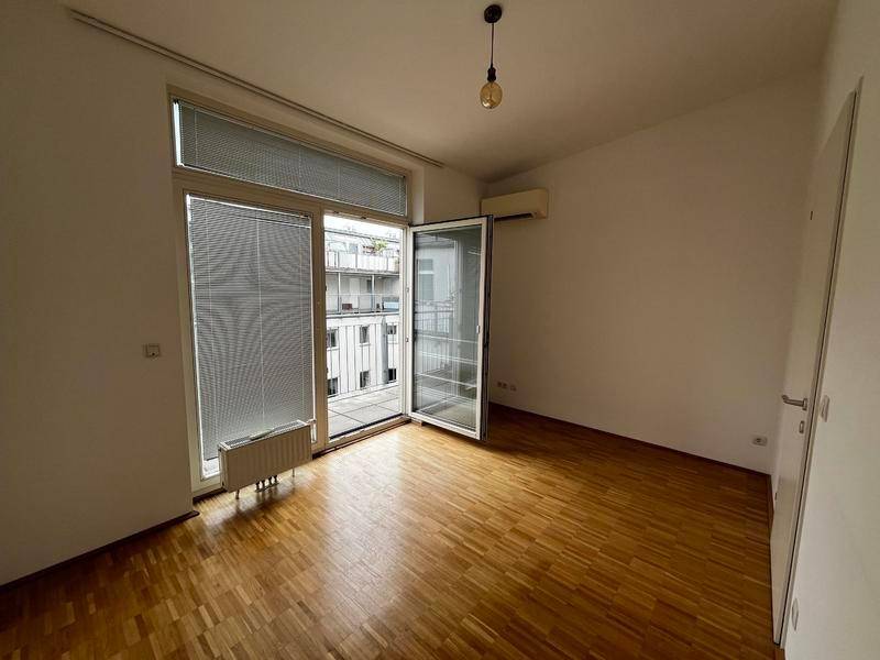 Helles Schlafzimmer mit Parkettboden, Klimaanlage und Zugang zu einem schmalen Balkon.