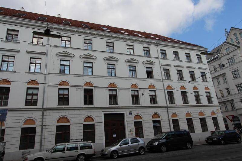 Klassische Fassade eines Mehrfamilienhauses mit vielen Fenstern und parkenden Autos davor.