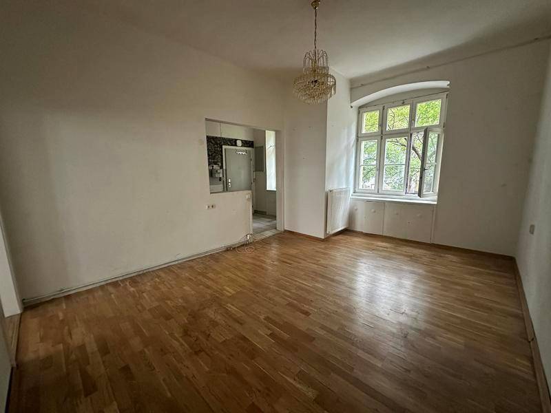 Geräumiges Zimmer mit Holzboden, Kronleuchter und Blick ins Grüne durch ein großes Fenster.