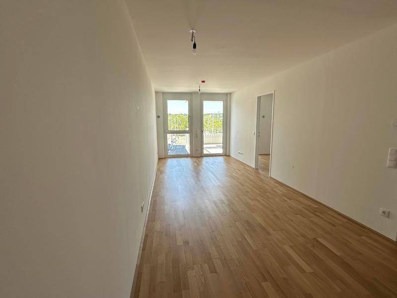 Helles, leeres Wohnzimmer mit Holzboden und Zugang zu einem Balkon.