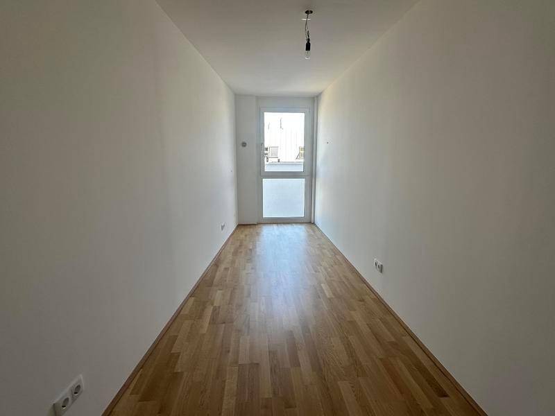 Schmaler, heller Raum mit Parkettboden und einer Tür, die zu einem Balkon führt.