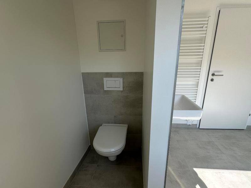 Separate Toilette mit Wand-WC und teilweiser Fliesenverkleidung in Grau.