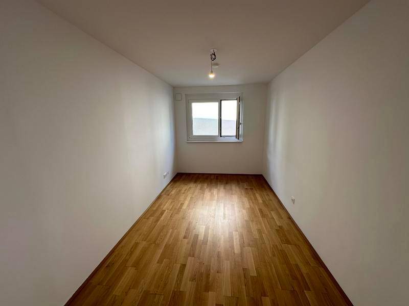 Leeres, helles Zimmer mit Holzfußboden und einem Fenster, ideal als Schlaf- oder Arbeitszimmer.