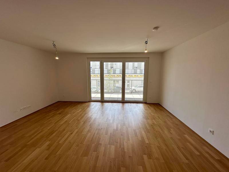Helles, leeres Wohnzimmer mit Holzfußboden und großen Schiebetüren zum Balkon.