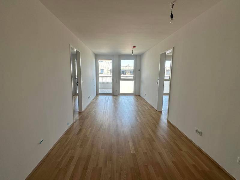 Geräumiger, leerer Wohnbereich mit hellem Parkettboden und Zugang zu einem Balkon.