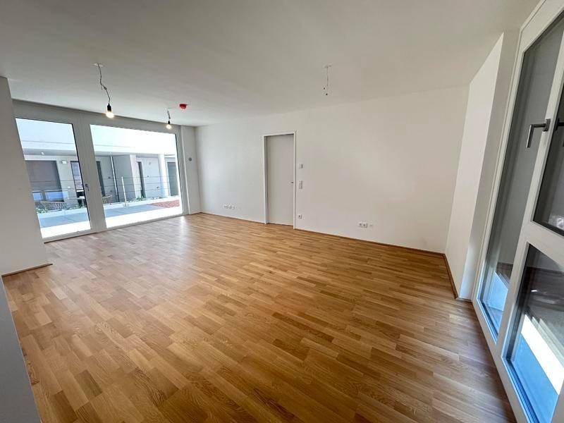 Heller, leerer Wohnraum mit großen Fenstern und hellem Holzparkettboden.