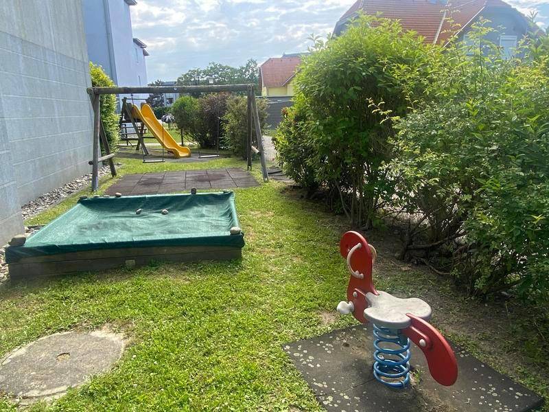 Gemeinschaftlicher Spielplatz mit Schaukeln, Rutsche und Sandkasten, umgeben von grünen Büschen.