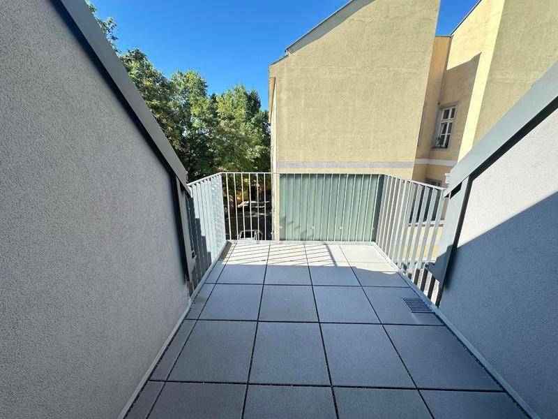 Schmaler Balkon mit Fliesenboden und Metallgeländer, bietet einen Blick auf Bäume und ein Nachbargebäude.
