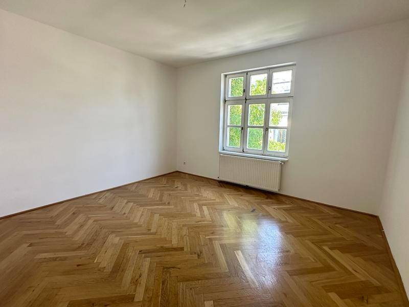 Leerer Raum mit hellem Fischgrätparkettboden und Fenster mit Blick ins Grüne.