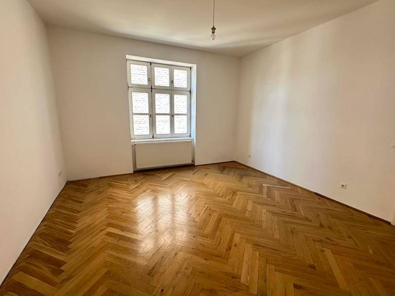 Geräumiger, leerer Raum mit klassischem Fischgrätparkett und großem Fenster.