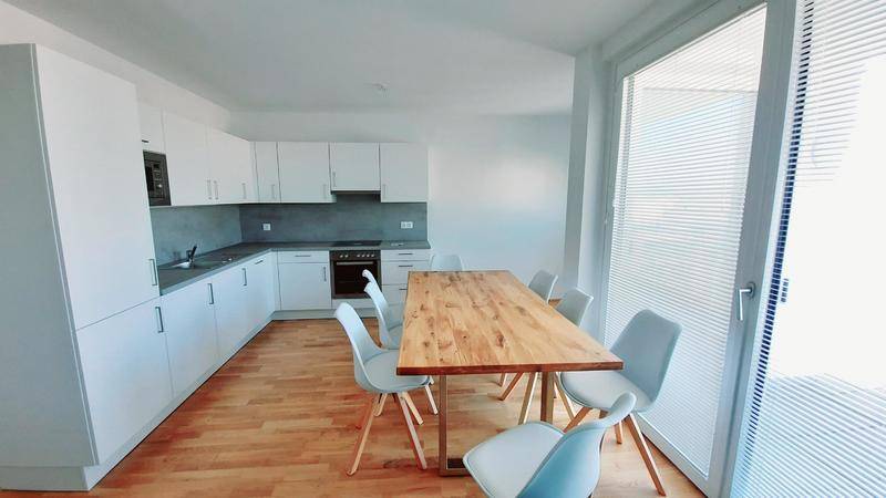 Moderne Einbauküche mit weißer Front, Holztisch und viel Tageslicht durch große Fenster.