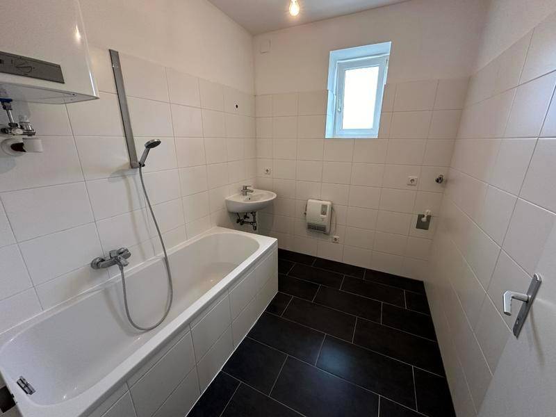 Helles Badezimmer mit Badewanne, Waschbecken und Fenster, weiß gefliest mit dunklem Boden.