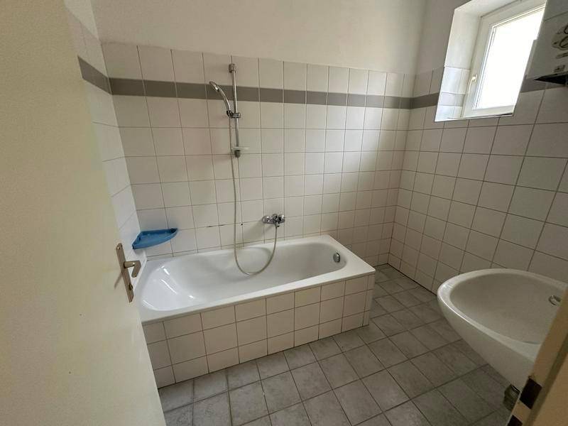 Badezimmer mit Badewanne, Waschbecken und Fenster, ausgestattet mit weißen Fliesen.