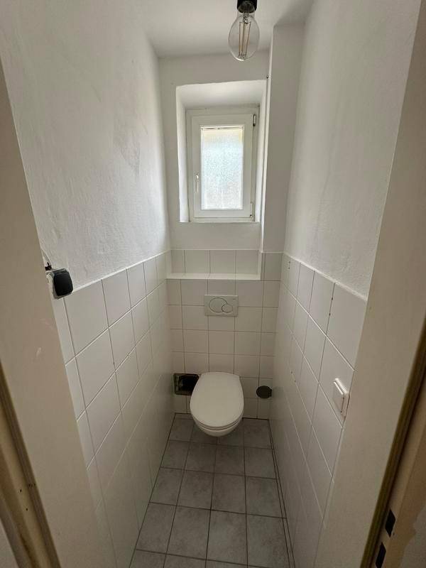 Kleines WC mit weiß gefliesten Wänden, Toilette und einem Fenster für Belüftung.