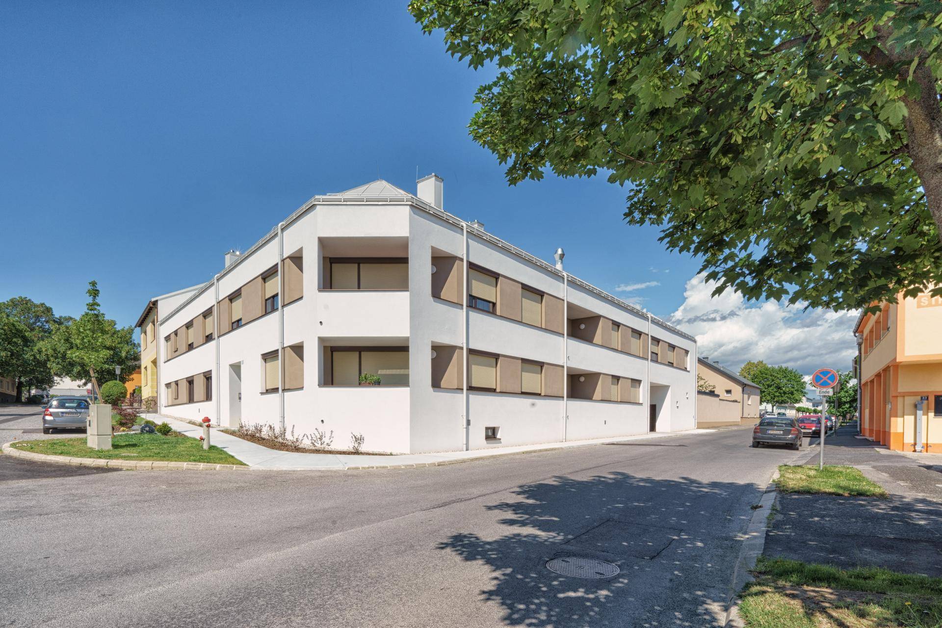 Modernes Wohngebäude an einer Straßenecke mit weißer Fassade und mehreren Balkonen unter blauem Himmel.