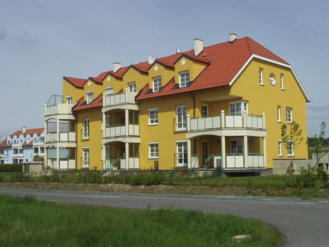 Langes Mehrfamilienhaus mit leuchtend gelber Fassade, roten Dächern und mehreren Balkonen in ländlicher Umgebung.