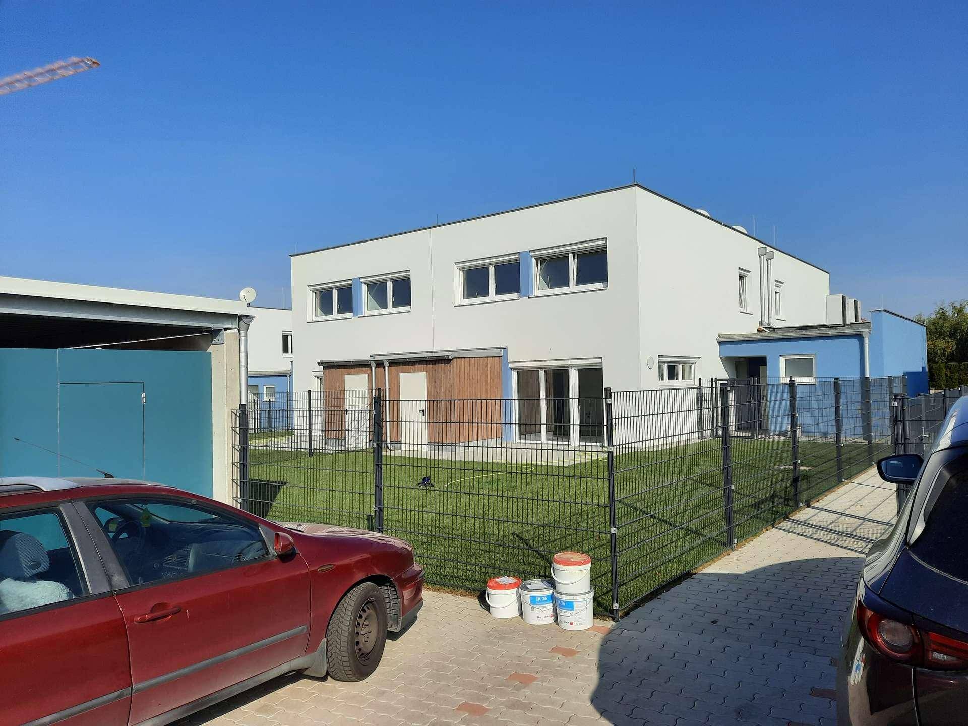 Moderne weiße Doppelhaushälfte mit blauen Akzenten und Gartenbereich, umzäunt, mit Fahrzeugen im Vordergrund.