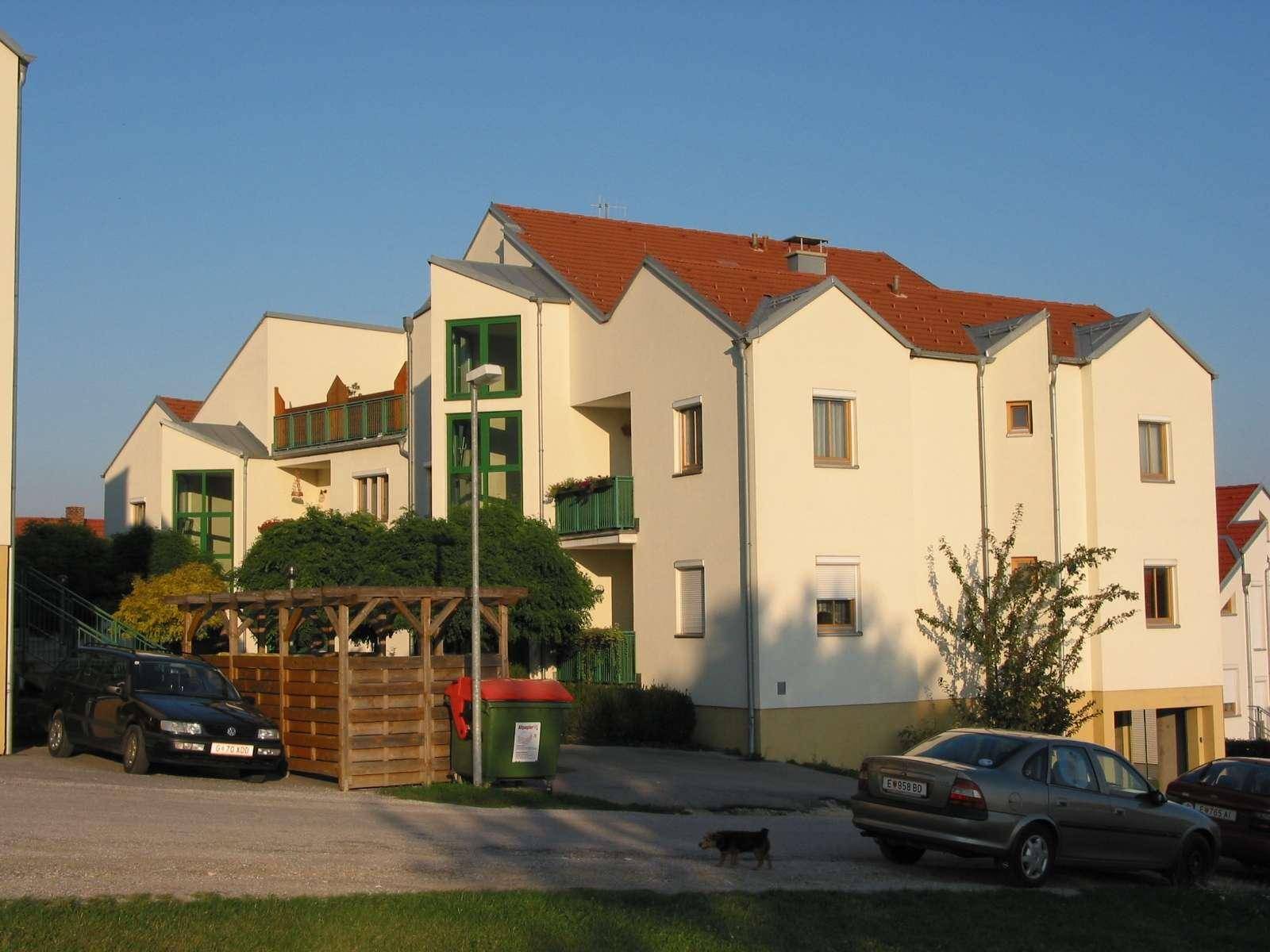 Wohngebäude mit heller Fassade, roten Dächern und Parkplätzen im Vordergrund, umgeben von Grünflächen.