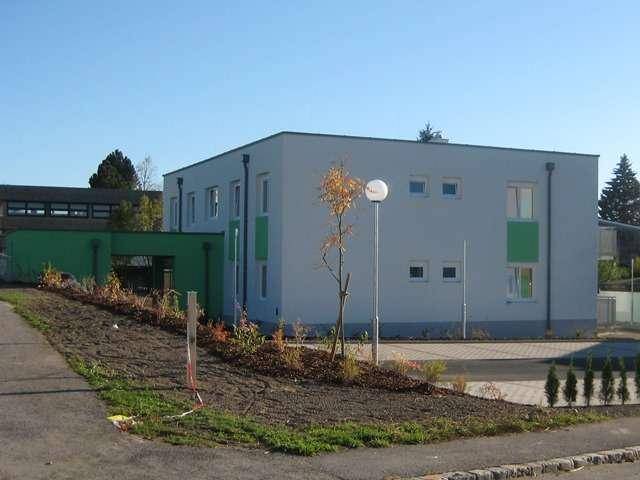 Modernes Gebäude mit grünen und grauen Fassadenbereichen sowie neu angelegten Grünflächen und Parkplätzen.