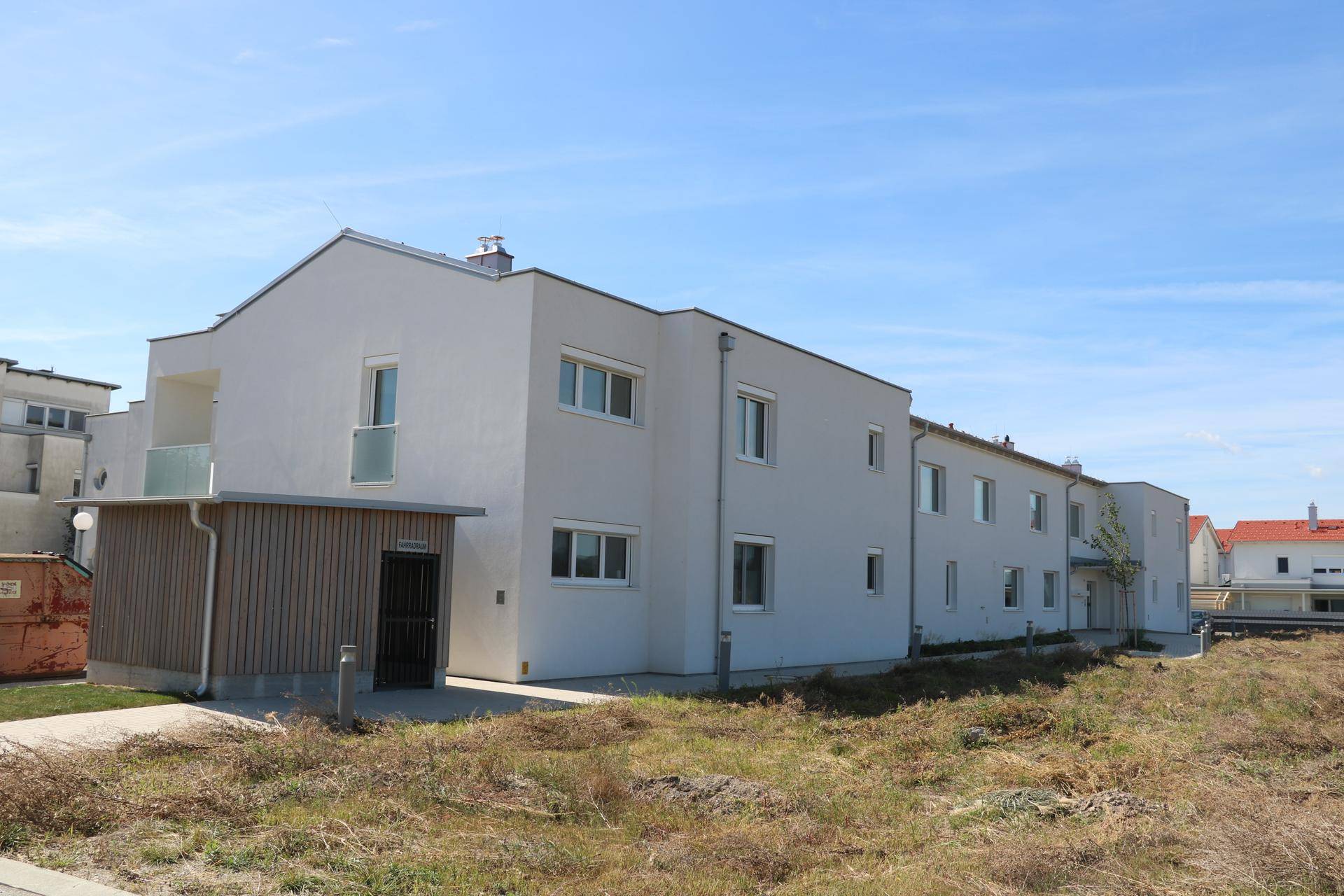 Modernes Mehrfamilienhaus mit heller Fassade, Balkonen und einem angrenzenden Holzschuppen in ländlicher Umgebung.