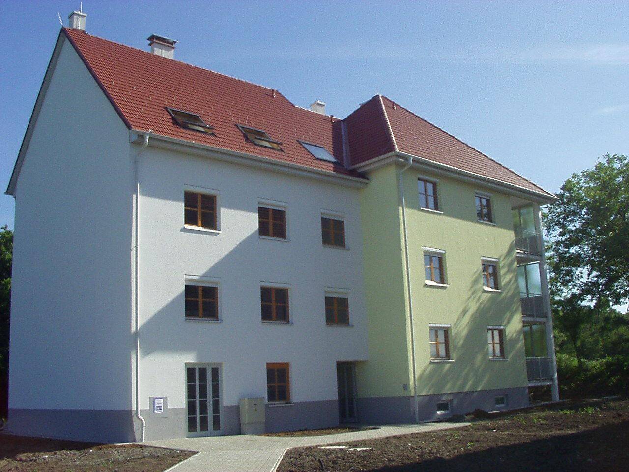 Neues Mehrfamilienhaus mit weißer und hellgrüner Fassade, rotem Satteldach und verglasten Balkonen.