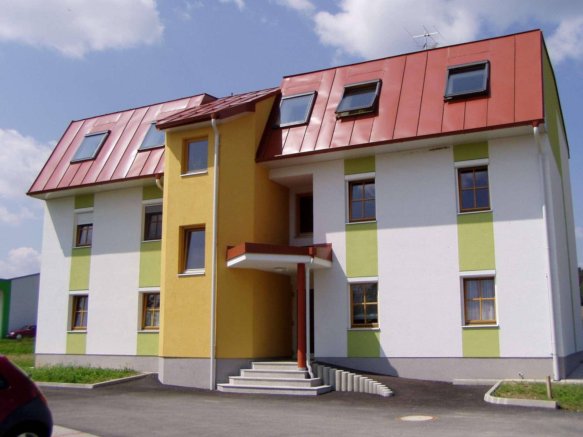 Neubau mit weißer, gelber und grüner Fassade, roten Dachziegeln und Rollläden an den Fenstern.