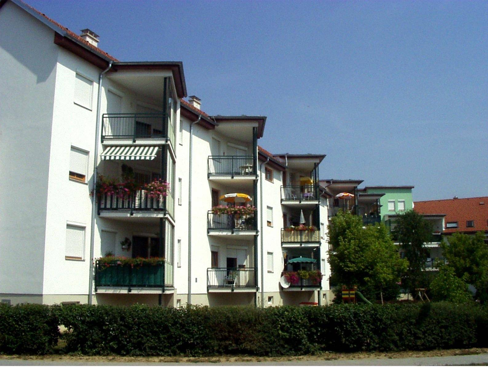 Modernes Mehrfamilienhaus mit weißen Fassaden, zahlreichen Balkonen, Markisen und Blumenkästen, umgeben von Grünflächen.