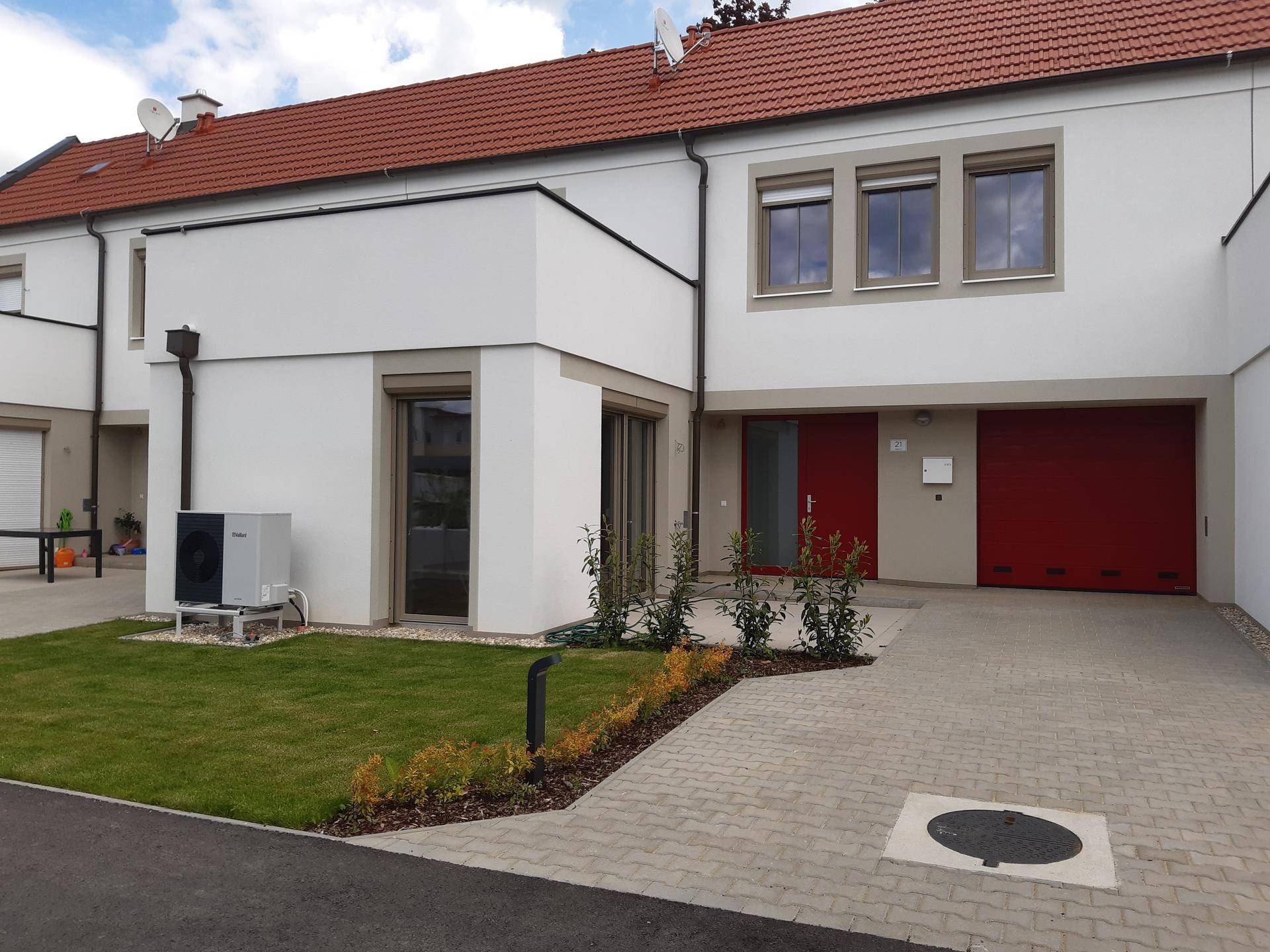 Modernes Reihenhaus mit roter Garagentür, kleinem Vorgarten und sichtbarer Wärmepumpe.