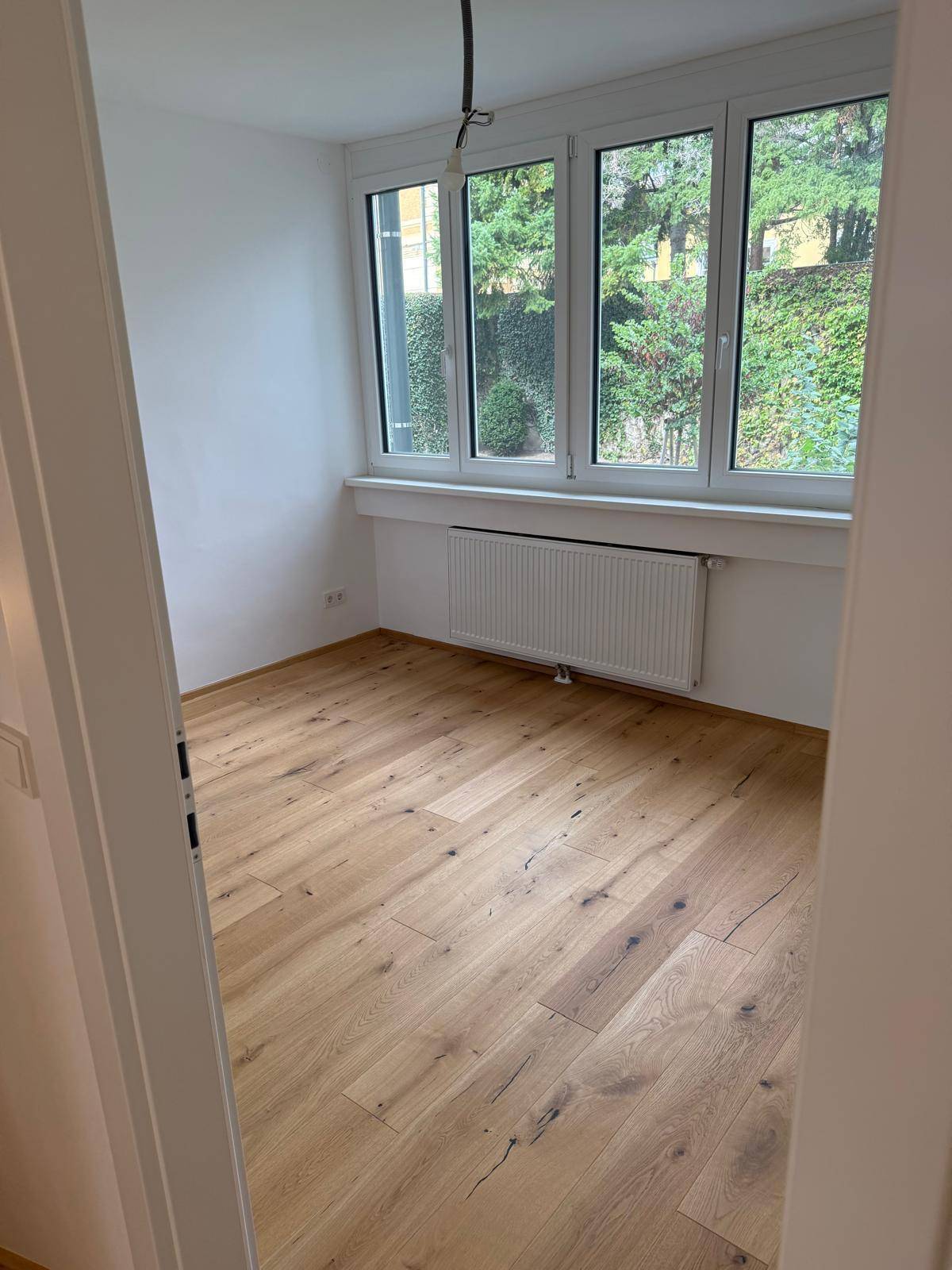 Helles Zimmer mit Blick ins Grüne, Holzboden und großen Fenstern.