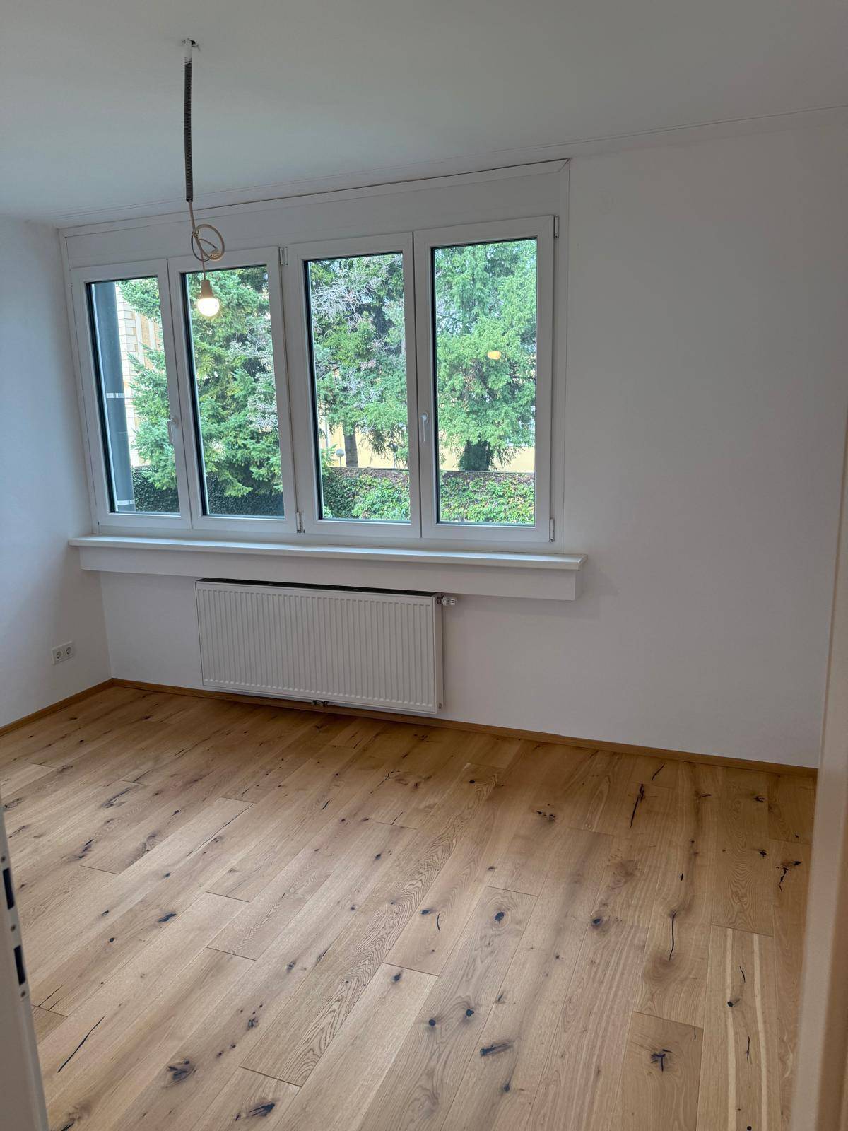 Geräumiges Zimmer mit Holzboden und Blick auf grüne Bäume durch die Fenster.