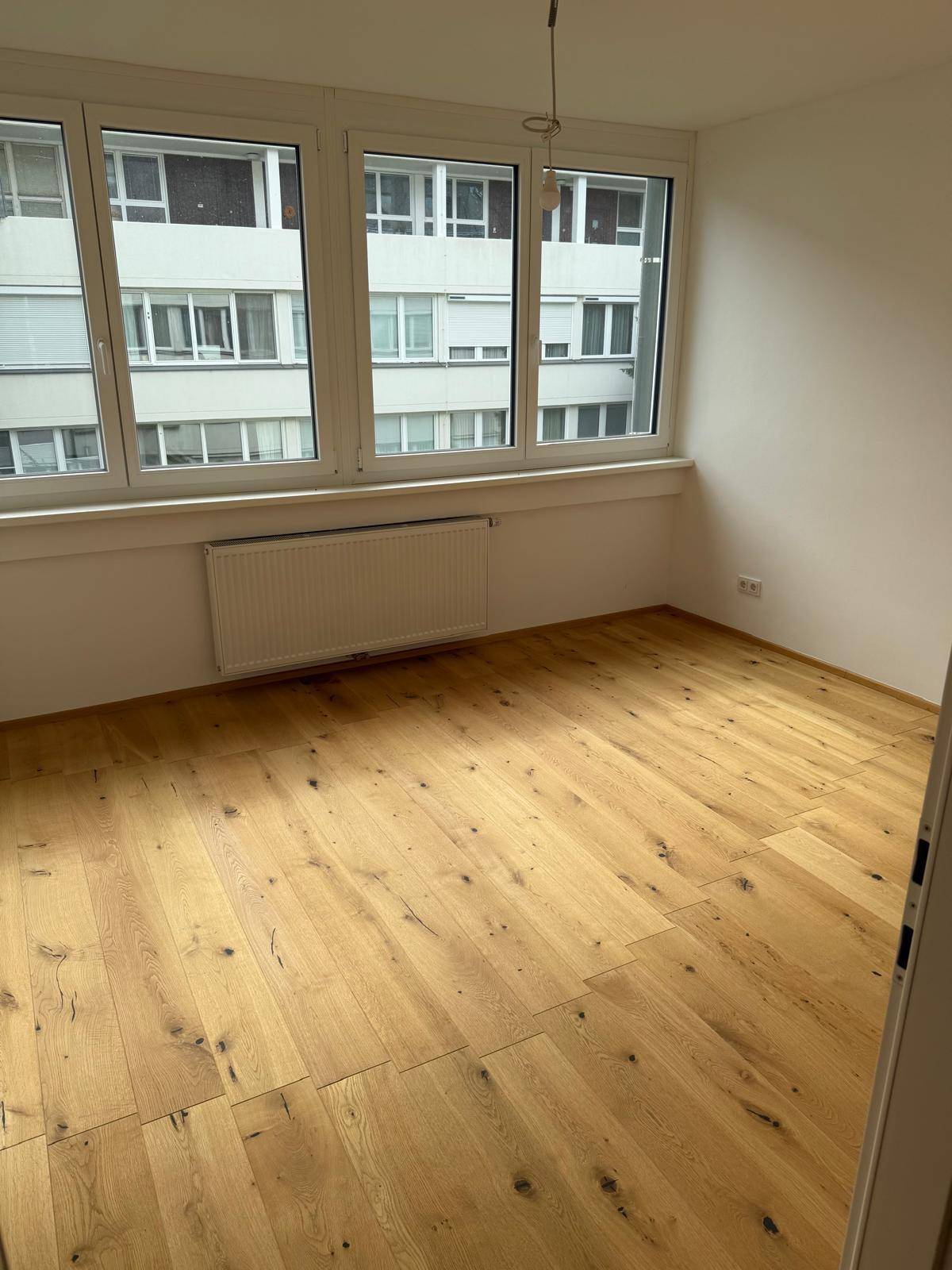 Helles Zimmer mit Holzboden und großen Fenstern, die viel Tageslicht hereinlassen.
