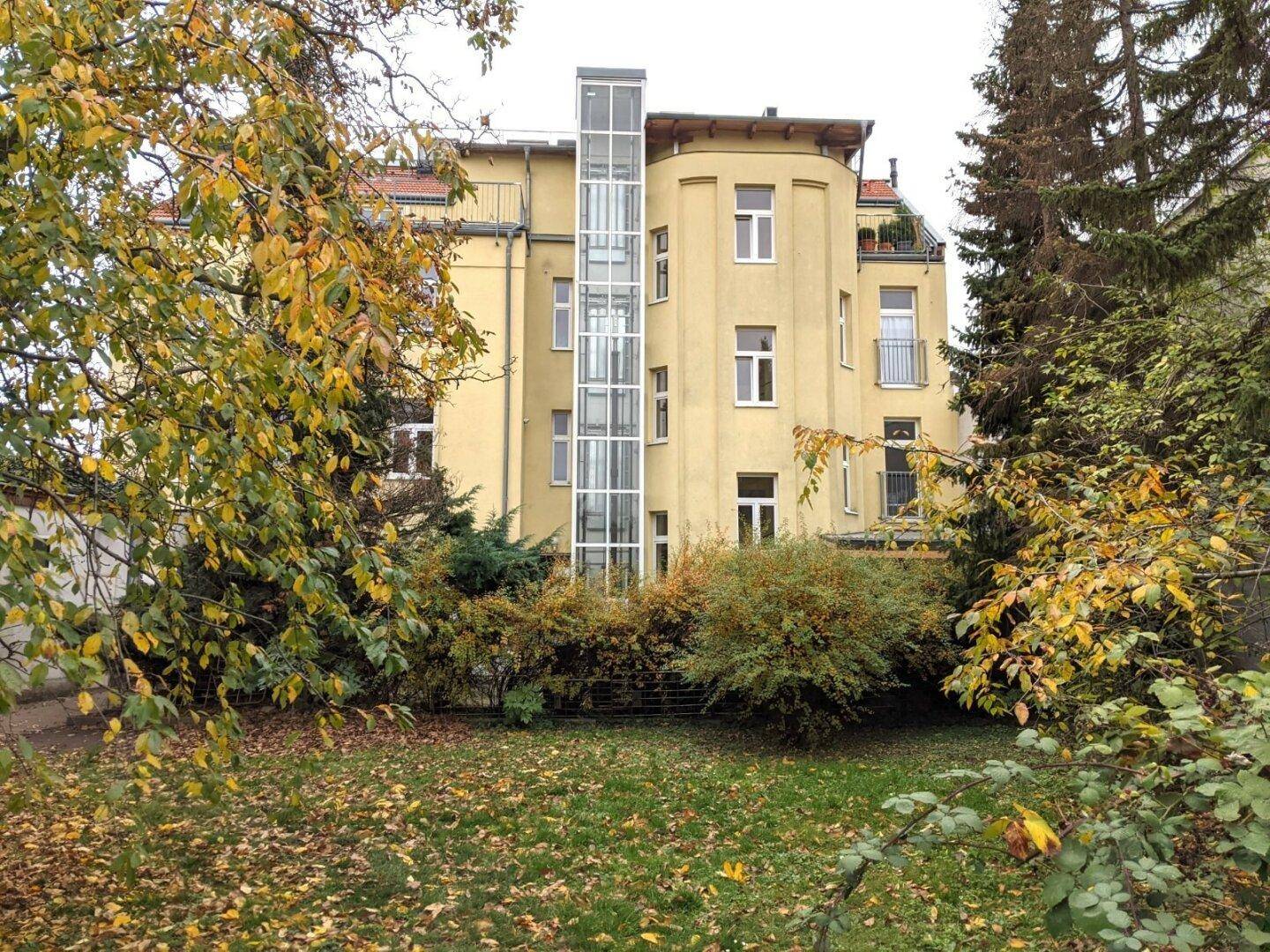 Rückseite eines gelben Gebäudes mit einem modernen gläsernen Außenaufzug, umgeben von Herbstbäumen.