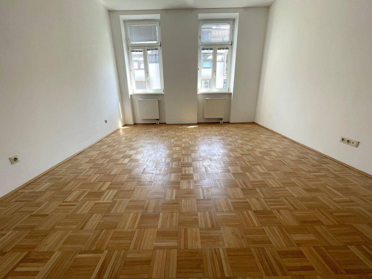 Helles, leeres Zimmer mit klassischem Parkettboden und zwei großen Fenstern für viel Tageslicht.