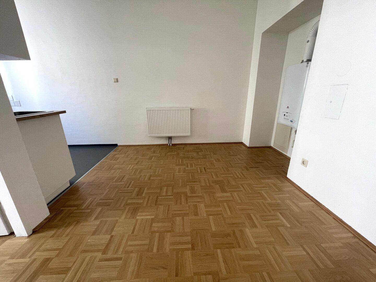 Heller Raum mit Parkettboden und Heizkörper, an der Wand ist ein Warmwasserbereiter sichtbar.